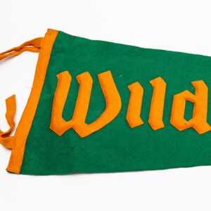 May include: Green felt pennant with orange trim and lettering. The word "Wildwood" is displayed in large, orange, stylized letters. The pennant has orange ties at the top.