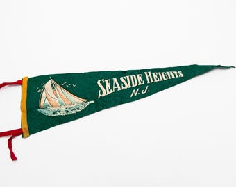 May include: A vintage green pennant with the words "SEASIDE HEIGHTS N.J." in white. A sailboat graphic is on the pennant. The pennant has a yellow border and red ties.