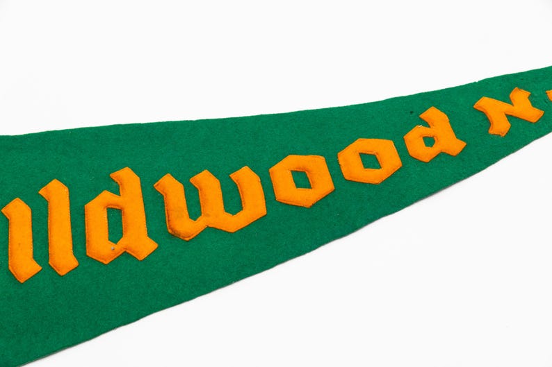 May include: Green felt pennant with orange lettering that reads "Wildwood N". The pennant is a classic shape, with a pointed end. The lettering is in a bold, blocky font.
