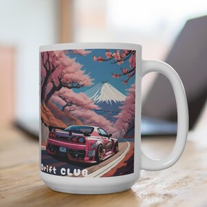 Könnte beinhalten: Weißer Keramikbecher mit einem Design eines pinkfarbenen Sportwagens auf einer Straße, vor dem Hintergrund von Kirschblütenbäumen und einem schneebedeckten Berg. Der Becher trägt den Text "Drift Club".