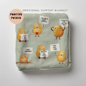 Peut inclure: Une couverture vert clair avec des illustrations de pommes de terre souriantes tenant des pancartes avec des messages positifs. La couverture porte les inscriptions "EMOTIONAL SUPPORT BLANKET" et "POSITIVE POTATO".