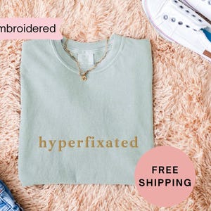 May include: A light green sweatshirt with the text "hyperfixated" embroidered in gold on the front.
