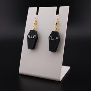 May include: A pair of black coffin-shaped earrings with gold-colored hooks. Each earring has the letters "R.I.P" in white. The earrings are displayed on a white stand against a black background. The earrings are a Halloween-themed accessory.