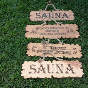 Op de afbeelding: Vier houten borden met touwhangers, elk met een unieke boodschap. De bovenste en onderste borden luiden "SAUNA". Het tweede bord zegt "GOOD PEOPLE DRINK GOOD BEER". Het derde bord zegt "STRESS FREE ZONE". De borden liggen op groen gras.