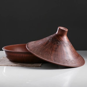 May include: A brown ceramic tagine pot with a conical lid, partially open to reveal the interior. The lid features an ornate, etched design. The pot sits on a neutral-colored surface, suggesting a culinary or decorative theme.
