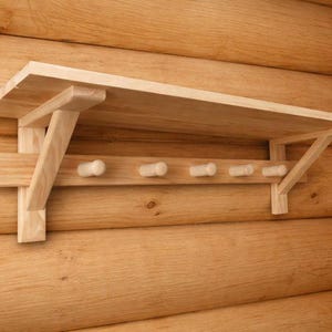 May include: A light-coloured wooden wall shelf with a row of pegs below. The shelf is supported by angled brackets and is mounted on a wooden wall. The pegs are cylindrical and evenly spaced.