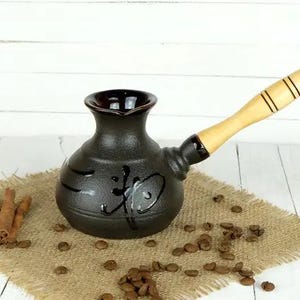 Op de afbeelding: Een zwarte keramische Turkse koffiepot met een houten handvat. De pot heeft een bolvormig lichaam en een smalle hals. De handgreep is lichtgekleurd hout met zwarte strepen. De pot is versierd met een zwart Aziatisch karakter.