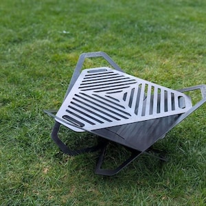Op de afbeelding: Een zwarte metalen vuurplaats met een driehoekig rooster. De vuurplaats staat op een groene grasmat.