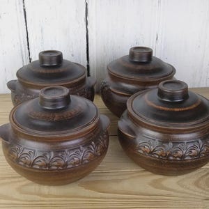 May include: Four brown ceramic pots with lids, arranged on a wooden surface. Each pot has a round shape with a lid and small handles. The pots have a decorative floral pattern around the base.