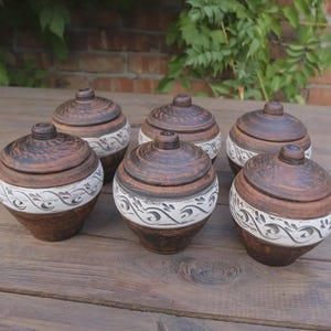 Puede incluir: Seis tarros de cerámica hechos a mano con tapas. Los tarros son marrones con una banda decorativa blanca con un patrón de remolino. Las tapas también son marrones con un pequeño pomo en la parte superior. Los tarros están dispuestos sobre una superficie de madera.