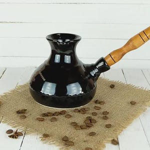 Könnte beinhalten: Eine schwarze Keramik-Kaffeekanne mit Holzgriff, auf einem Jutegewebe. Die Kanne hat eine glänzende Oberfläche und ist von Kaffeebohnen und Zimtstangen umgeben. Das Wort "espresso" ist auf der Seite aufgedruckt.