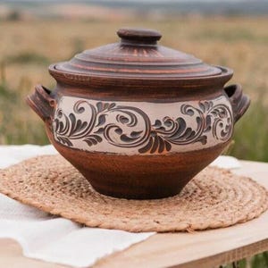 Può includere: Un vaso di ceramica marrone con un motivo floreale bianco. Il vaso ha due manici e un coperchio. È seduto su un tappetino intrecciato.