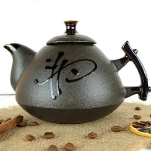 Könnte beinhalten: Eine dunkelgraue Keramik-Teekanne mit Deckel und gebogenem Griff. Die Teekanne hat schwarze Kalligraphie auf der Vorderseite. Sie steht auf einer Jutefläche mit Zimtstangen, Kaffeebohnen und einer getrockneten Zitronenscheibe.