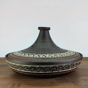 Puede incluir: Una olla de tagine de cerámica marrón y negra con tapa cónica. La tapa y la base presentan patrones decorativos blancos y marrones. El tagine está diseñado para cocinar y servir platos marroquíes y del norte de África. Se encuentra sobre una superficie de madera.