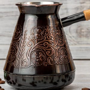 Puede incluir: Una cafetera metálica oscura con grabados florales y figurativos de color cobre ornamentados. La cafetera tiene un mango largo con un agarre de madera y está rodeada de granos de café. El diseño de la cafetera incluye los números "19.11-20.11" y "23.VII.22".
