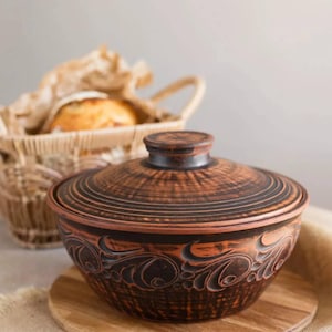 May include: A brown ceramic pot with a lid, decorated with a floral pattern. The pot is sitting on a wooden base and is in front of a wicker basket.