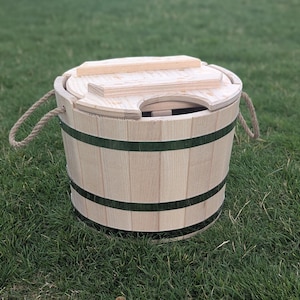 Puede incluir: Un barril de madera de color marrón claro con bandas verdes oscuras y asas de cuerda. El barril tiene una tapa de madera y está sobre hierba verde. Este artículo es adecuado para almacenamiento o fines decorativos.