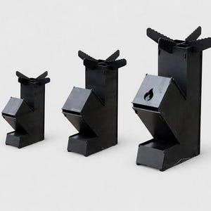 Pode incluir: Três fogões de campismo de metal preto de tamanhos variados são exibidos contra um fundo branco. Os fogões têm uma base retangular, uma frente angulada e um topo com quatro pinos. O fogão maior tem um recorte de chama.