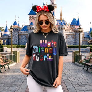 Puede incluir: Camiseta negra con las palabras "Walt Disney World" en texto colorido y estilizado. La persona lleva una diadema con orejas de Minnie Mouse con un lazo rojo y gafas de sol. El fondo presenta un castillo y un parque.