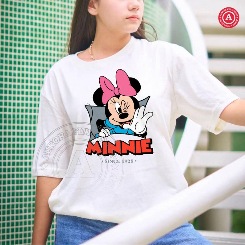 May include: White t-shirt featuring a Minnie Mouse graphic. The graphic shows Minnie Mouse with a pink bow, waving, and the word "MINNIE" in red block letters. The text "SINCE 1928" is below the name.