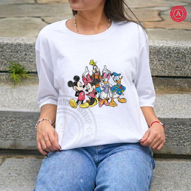 May include: White t-shirt with a colourful graphic of Mickey Mouse, Minnie Mouse, Donald Duck, Daisy Duck, and Goofy. The shirt has short sleeves and a crew neck. The characters are in their classic outfits. The shirt is worn by a person.