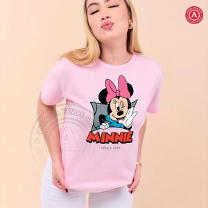 May include: A light pink t-shirt featuring a graphic of Minnie Mouse waving. Minnie is wearing a pink bow, blue top, and white gloves. The word "MINNIE" is in red letters with a blue outline, and the text "SINCE 1928" is below.