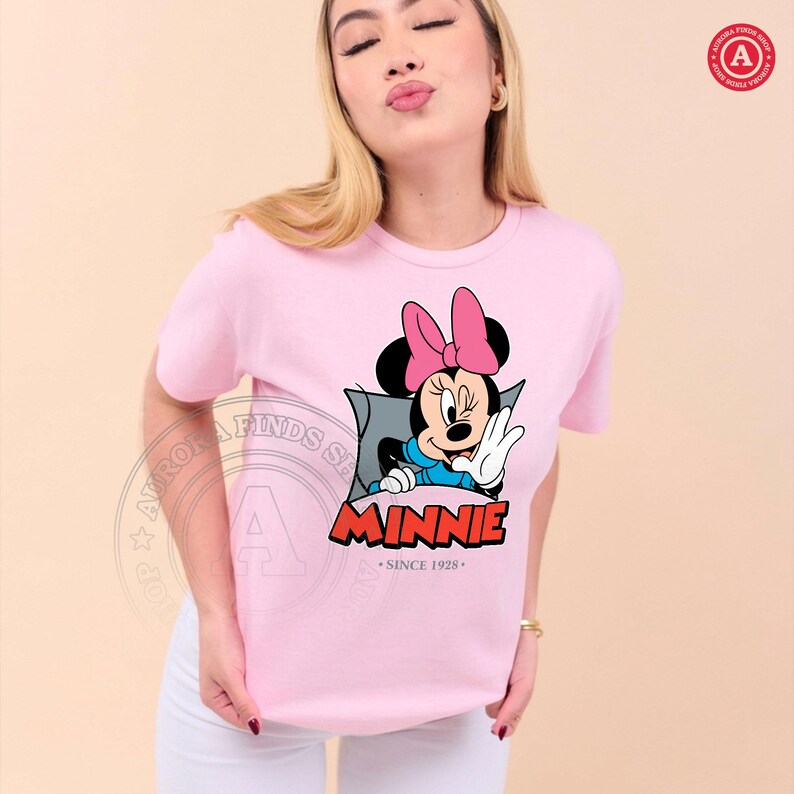 May include: A light pink t-shirt featuring a graphic of Minnie Mouse waving. Minnie is wearing a pink bow, blue top, and white gloves. The word "MINNIE" is in red letters with a blue outline, and the text "SINCE 1928" is below.
