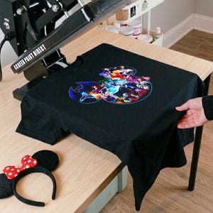 K&ouml;nnte beinhalten: Schwarzes T-Shirt mit einem farbenfrohen Grafikdesign mit verschiedenen Zeichentrickfiguren. Ein Minnie-Mouse-Ohren-Haarreif mit einer rot-wei&szlig; gepunkteten Schleife befindet sich neben dem Shirt. Das Shirt liegt auf einem Holztisch.