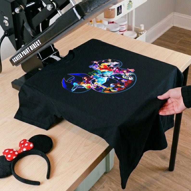 K&ouml;nnte beinhalten: Schwarzes T-Shirt mit einem farbenfrohen Grafikdesign mit verschiedenen Zeichentrickfiguren. Ein Minnie-Mouse-Ohren-Haarreif mit einer rot-wei&szlig; gepunkteten Schleife befindet sich neben dem Shirt. Das Shirt liegt auf einem Holztisch.
