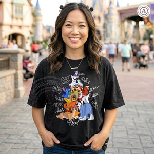 K&ouml;nnte beinhalten: Schwarzes T-Shirt mit einer bunten Grafik von Zeichentrickhunden, darunter Bolt, Stitch und Pluto, mit ihren Namen. Das Shirt hat einen Rundhalsausschnitt und kurze &Auml;rmel. Das Design enth&auml;lt den Text "Aurora Finds".