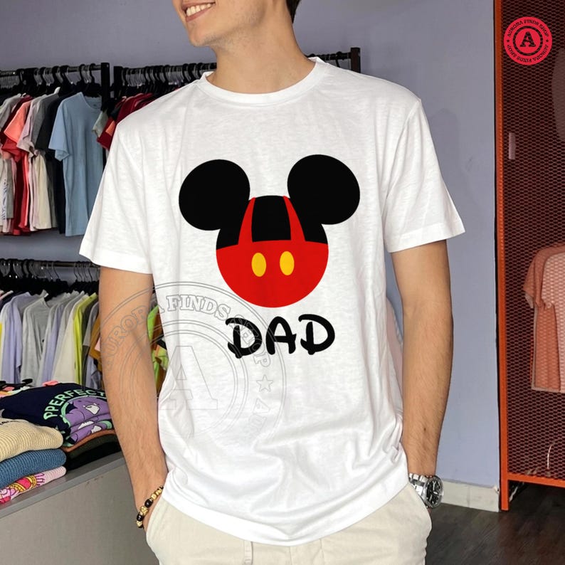 May include: White t-shirt featuring a Mickey Mouse-inspired design with black ears, a red face, and yellow dots. The word "DAD" is printed in black below the graphic. The shirt is short-sleeved.