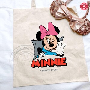 May include: Beige canvas tote bag featuring a Minnie Mouse graphic with a pink bow and the word "MINNIE" in red. The text "SINCE 1928" is below the name. A pair of sequined Minnie Mouse ears with a pink bow are also in the image.