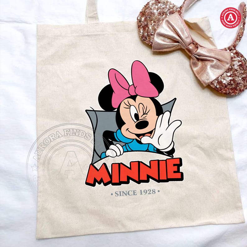 May include: Beige canvas tote bag featuring a Minnie Mouse graphic with a pink bow and the word "MINNIE" in red. The text "SINCE 1928" is below the name. A pair of sequined Minnie Mouse ears with a pink bow are also in the image.