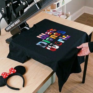 Puede incluir: Camiseta negra con las palabras "Walt Disney World" en texto colorido y estilizado. Una diadema de orejas de Minnie Mouse con un lazo rojo y blanco con lunares est&aacute; sobre la mesa. La camiseta est&aacute; sobre una mesa de madera.