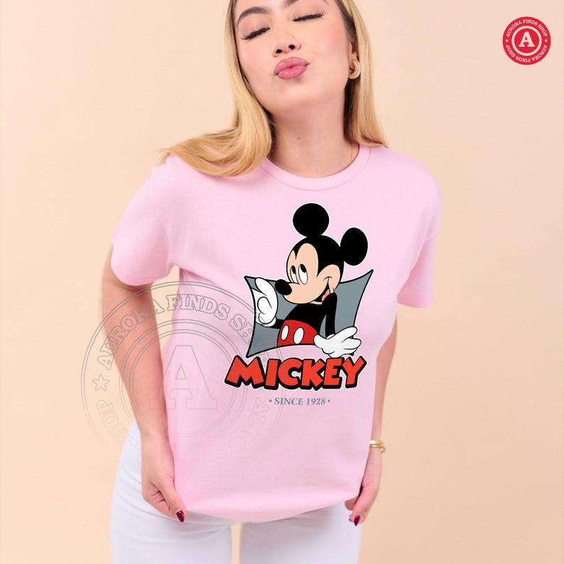 May include: A light pink t-shirt featuring a graphic of Mickey Mouse pointing. The graphic includes the word "Mickey" in red letters and the text "Since 1928". The shirt is a crew neck style.