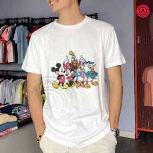 May include: White t-shirt with a vibrant graphic of Disney characters including Mickey and Minnie Mouse, Goofy, Daisy Duck, and Donald Duck. The tee has a crew neck and short sleeves. The image shows a person wearing the shirt.