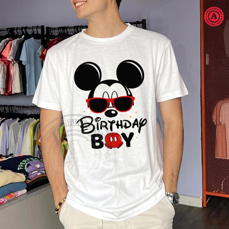 May include: White t-shirt featuring a graphic of Mickey Mouse wearing red sunglasses. The design includes the text "Birthday Boy" in black and red. The shirt is short-sleeved and appears to be made of cotton.