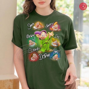 K&ouml;nnte beinhalten: Dunkelgr&uuml;nes T-Shirt mit den sieben Zwergen aus Schneewittchen. Jeder Zwerg ist mit seinem Namen in einer skurrilen Schriftart dargestellt. Der zentrale Zwerg ist Dopey in einem gr&uuml;nen Outfit.