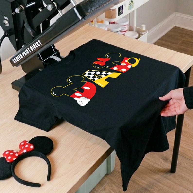 May include: Black t-shirt with a colorful graphic design featuring the word "DAD" in a Mickey Mouse-inspired font. The design incorporates red, yellow, and black elements. A Minnie Mouse ear headband with a red bow is also visible.