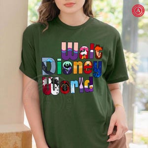 Puede incluir: Camiseta verde oscuro con la frase "Walt Disney World" en letras coloridas y estilizadas. Las letras incorporan varios dise&ntilde;os de personajes de Disney. La camiseta es de cuello redondo y manga corta.