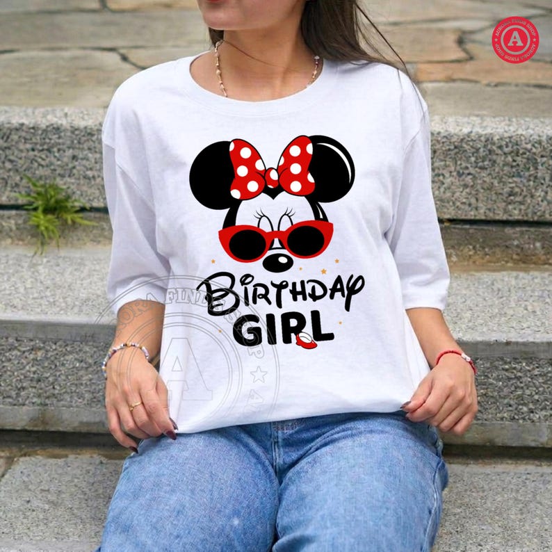 May include: White t-shirt featuring a Minnie Mouse design with a red bow and sunglasses. The text "Birthday Girl" is printed below the graphic in black. The shirt has three-quarter sleeves and is paired with blue jeans.