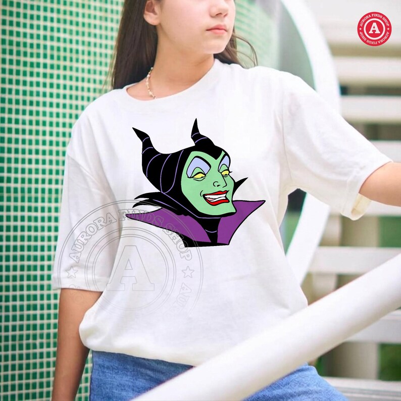 May include: White t-shirt featuring a cartoon graphic of Maleficent with a green face, black horns, and a purple collar. The design is centered on the shirt. The shirt has short sleeves.