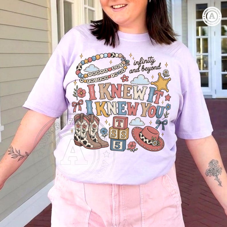Op de afbeelding: Een lichtpaars t-shirt met een grafische print met de tekst "I KNEW IT I KNEW YOU", "infinity and beyond", "A FRIEND IN ME" en "YOU'VE GOT". Het ontwerp bevat een kralenarmband, cowboylaarzen, een hoed en gestapelde blokken met letters.