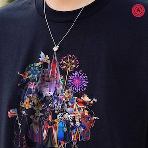 May include: Navy blue t-shirt featuring a colorful graphic of a castle, fireworks, and various cartoon characters. A silver necklace with a heart pendant is visible. The shirt has a round neck and short sleeves. The graphic includes characters from popular animated films.