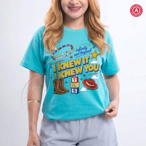 Puede incluir: Una camiseta de manga corta de color turquesa brillante con un estampado gr&aacute;fico que incluye el texto "I KNEW IT I KNEW YOU" y "infinity and beyond". El dise&ntilde;o muestra ilustraciones de botas vaqueras marrones, un sombrero rojo y bloques.