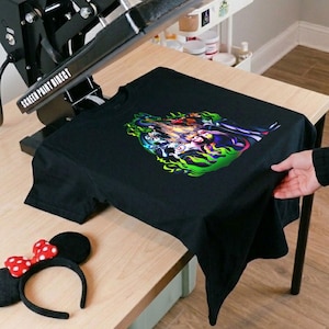 Puede incluir: Camiseta negra con un dise&ntilde;o gr&aacute;fico colorido con personajes en un dise&ntilde;o de llama verde y amarilla. Una prensa de calor negra est&aacute; encima de la camiseta. Una diadema de orejas de Minnie Mouse con un lazo de lunares rojos y blancos est&aacute; sobre la mesa.