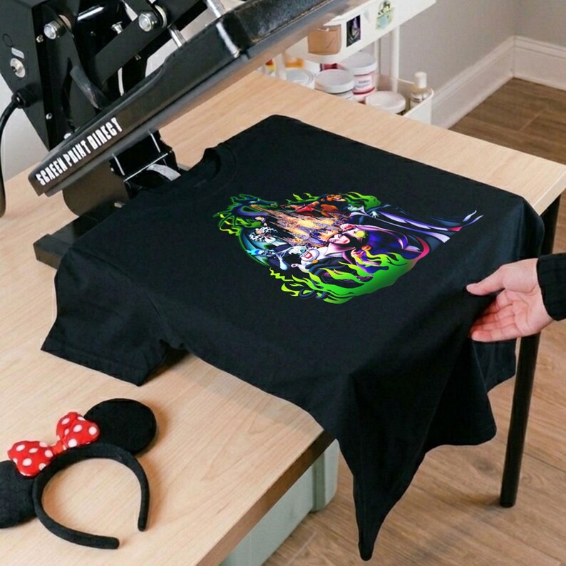 Puede incluir: Camiseta negra con un dise&ntilde;o gr&aacute;fico colorido con personajes en un dise&ntilde;o de llama verde y amarilla. Una prensa de calor negra est&aacute; encima de la camiseta. Una diadema de orejas de Minnie Mouse con un lazo de lunares rojos y blancos est&aacute; sobre la mesa.
