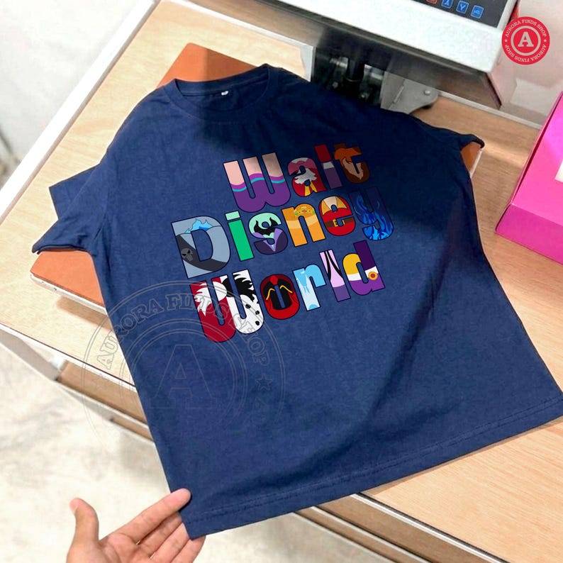 Puede incluir: Camiseta azul marino con las palabras "Walt Disney World" en letras coloridas y estilizadas. El dise&ntilde;o incorpora elementos de personajes de Disney dentro de las letras. La camiseta es de un material suave.