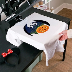 May include: White t-shirt featuring a Halloween-themed graphic. The design includes a jack-o'-lantern, a bat-like creature, and a crescent moon. The shirt is being pressed with a heat press machine. A Minnie Mouse ear headband is on the table.