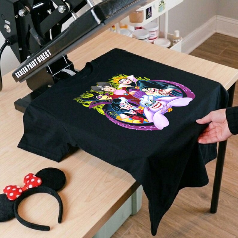 May include: Black t-shirt featuring a colorful graphic of cartoon villains. The design includes characters with various skin tones and hair colors, surrounded by flames and tentacles. A Minnie Mouse ear headband with a red bow is also visible.
